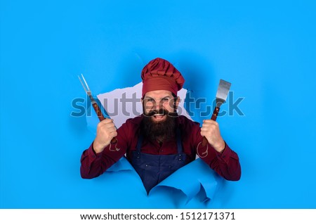 Grill cook. Chef man with BBQ cooking tools. Barbecue chef looking through paper hole. Picnic&barbecue. Cooking meat in park. Barbecue master. Chief cook in workwear with utensils for barbecue grill. Royalty-Free Stock Photo #1512171371