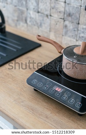 Vertical photo of kitchenware pan at small electric stove with control panel standing in modern kitchen. Marble wall tile and wooden surface table on blurred background