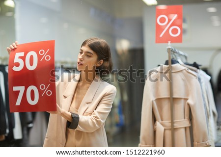 Waist up portrait of beautiful woman hanging red SALE signs on window display in clothes store, copy space