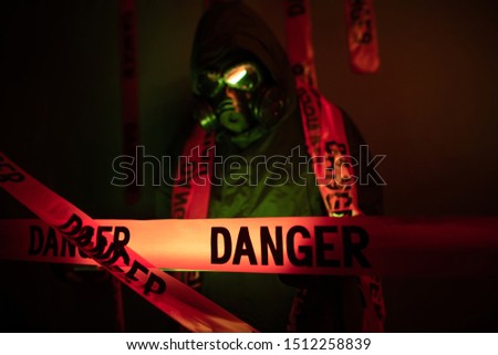 Portrait of a guy in a green protective suit with green rubber gloves with a gas mask on his face and a hood on his head standing near a green wall with a large number of danger tapes with a flashligh