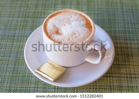 a Cup of frothy cappuccino on a saucer with a square piece of chocolate, on a uniform background of bamboo.