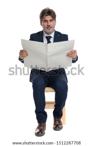elegant formal business man with navy suit sitting with newspaper on hands and looking to the camera serious on white studio background