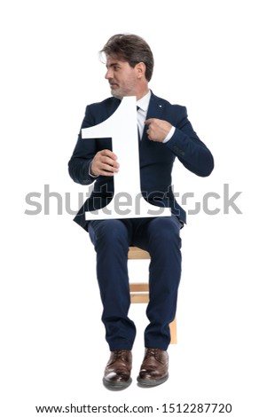 cocky formal business man with navy suit is sitting and holding a number one on one hand while pointing to himself and looking sideways confident on white studio background