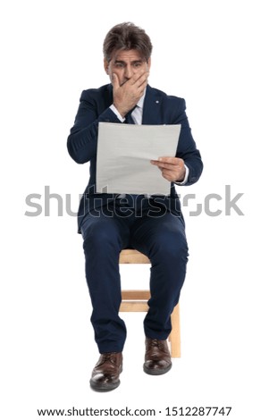 gorgeous formal business man with navy suit is sitting and reading the newspaper terrified on white studio background 