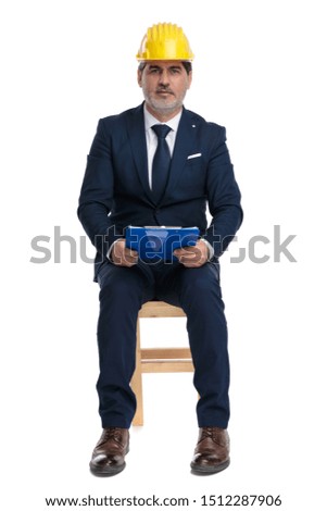 middle aged formal business man with navy suit and safety helmet is sitting and holding a clipboard on his hands serious on white studio background