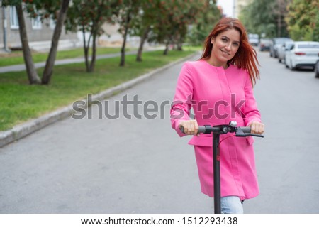 A red-haired young woman in ripped jeans and high-heeled sandals rides an electric scooter on the road. A girl in a pink jacket quickly moves around the city on a convenient electric transport.