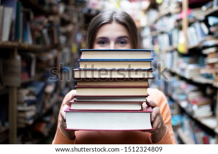 young girl student holds a stack of books in the library, she searches for literature and offers to read, a woman prepares for study, bookseller on the background of the bookstore, knowledge is power Royalty-Free Stock Photo #1512324809