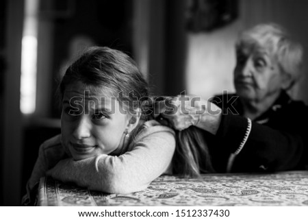 Grandmother braids her hair to a disgruntled little girl. Black and white picture.