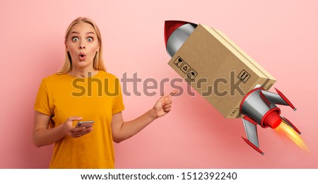 Blonde cute girl receives a priority fast box, like a rocket, from online shop order. Surprised and amazed expression. Pink background