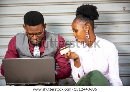 two young business couple sitting outdoors paying bills with credit card using a laptop while smiling