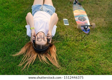 Young girl enjoy lay in green lawn and listening music after riding on her logboard.