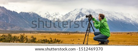 Photographer man taking nature photography with professional SLR camera at mountain landscape, New Zealand Banner. Tourist on travel adventure holiday shooting video on tripod panorama.