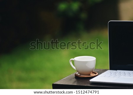 The picture of white laptop, a coffee cup above wooden table on blurred green grass background with copyspace. Selective focus