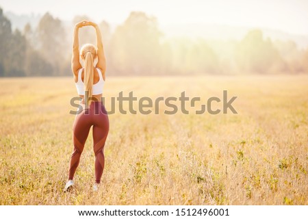Young woman blonde in red pants and white T-shirt performs warm before running in park.