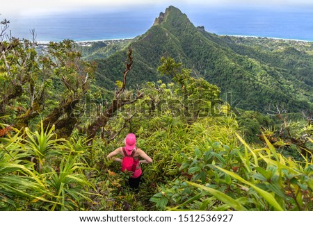 Rear view of a blonde young woman hiker with backpack standing on the top of exotic mountain and enjoying stunning valley view on Te Manga track in Rarotonga, Cook Islands.