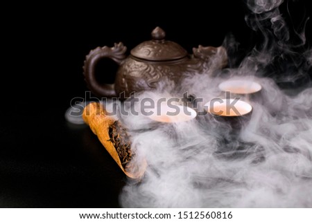 Ceramic tea cups, clay tea pot and dried black tea in a scoop with vapor on a black background.