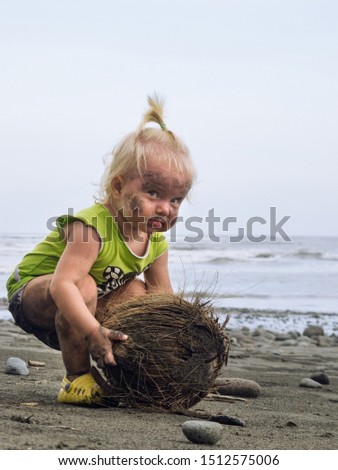 Funny portrait of smiling child with dirty face and hands playing with coconut. Having fun on black sand sea beach. Family travel lifestyle, activity on summer vacation with baby on tropical island.