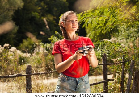 Beautiful caucasian brunette girl holding a camera, standing in the field by a wooden fence in the countryside, wanderlust, hike or picnic in the nature concept 