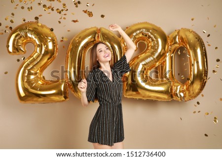 Excited young woman near golden 2020 balloons on beige background. New Year celebration