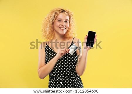Caucasian young woman's half-length portrait on yellow studio background. Beautiful female model in black dress. Concept of human emotions, facial expression. Showing phone screen and payment card.