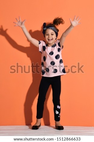 Laughing kid girl in is standing holding her hands up, jumping, funny dancing, having fun, showing ten on orange background