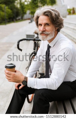 Photo of serious mature businessman in formal clothes drinking coffee takeaway while sitting on bench with bicycle outdoors