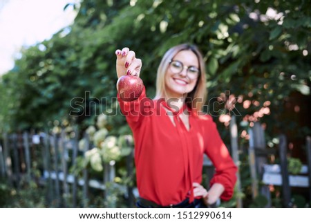 Young blond woman, wearing eyeglasses and red blouse, holding red claret apple in her hand. Close-up picture of female student with blurred face in the garden. An apple a day keeps doctor away.
