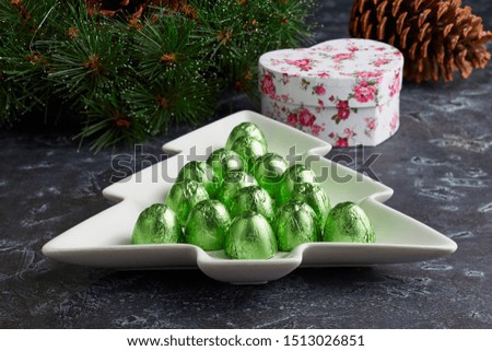 Plate of chocolates wrapped in green foil on the background of a Christmas wreath, a big cone and a gift box.