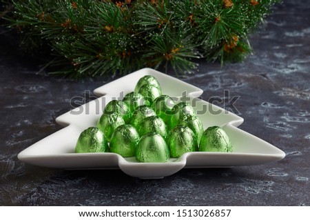 Plate in the shape of a Christmas tree with candy in green foil wrappers on the background of a Christmas wreath.