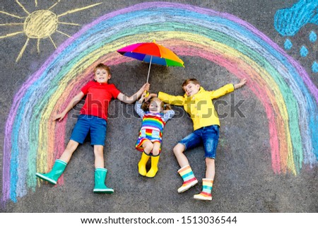 three little children, two school kids boys and toddler girl having fun with with rainbow picture drawing with colorful chalks on asphalt. Siblings in rubber boots painting on ground playing together. Royalty-Free Stock Photo #1513036544