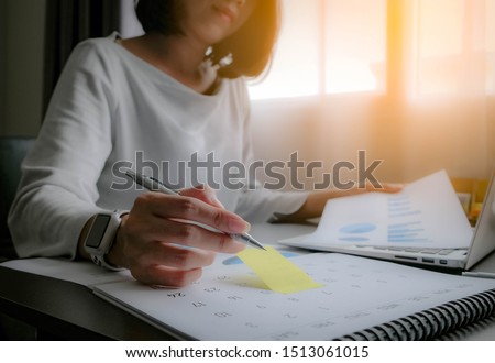 Asian woman is writing on yellow paper notes and put a warning on calendar. The other hand hold the report graph and has laptop on the front. Maybe preparing data for marketing report. window light Royalty-Free Stock Photo #1513061015