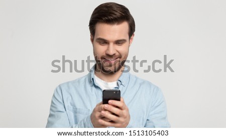 Smiling young man standing with smartphone in hands, looking at screen, reading pleasant message from beloved woman or friend, typing sms, playing online game, using apps isolated on grey background.