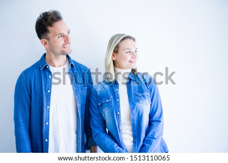 Young beautiful couple wearing denim shirt standing over isolated white background looking away to side with smile on face, natural expression. Laughing confident.