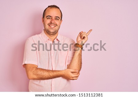 Young man wearing elegant shirt standing over isolated pink background with a big smile on face, pointing with hand and finger to the side looking at the camera.