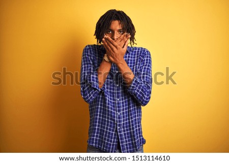 Afro man with dreadlocks wearing casual shirt standing over isolated yellow background shocked covering mouth with hands for mistake. Secret concept.