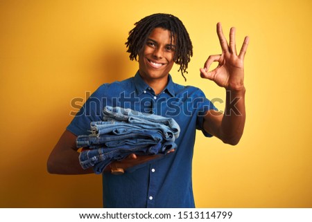 Afro man with dreadlocks holding stack of folded jeans over isolated yellow background doing ok sign with fingers, excellent symbol