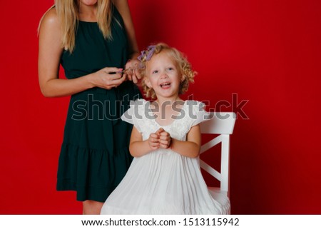 Mother curls daughter hair. Little blonde girl sitting on a white chair isolated on red