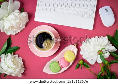 Fashionable feminine workspace flat lay. Female home office with laptop, coffee cup, peony flowers, macarons cake on pink background. Femininity background. Top view