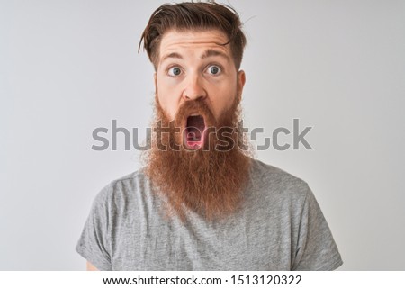 Young redhead irish man wearing t-shirt standing over isolated grey background scared in shock with a surprise face, afraid and excited with fear expression