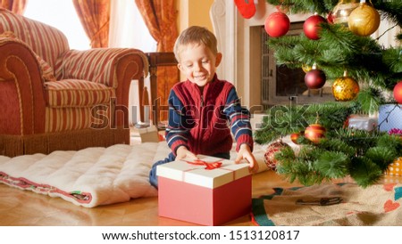 Cheerful laughing toddler boy opening Christmas gift box while sitting on the floor under Christmas tree