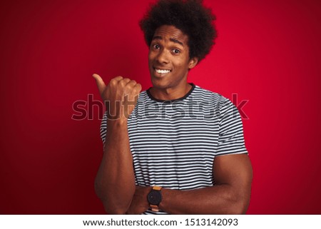 Young american man with afro hair wearing navy striped t-shirt over isolated red background smiling with happy face looking and pointing to the side with thumb up.