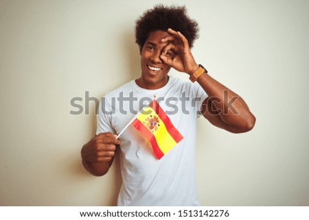 Young afro american man holding Spain Spanish flag standing over isolated white background with happy face smiling doing ok sign with hand on eye looking through fingers