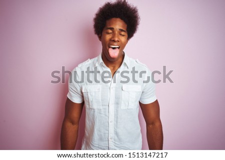 Young american man with afro hair wearing white shirt standing over isolated pink background sticking tongue out happy with funny expression. Emotion concept.