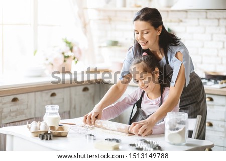 Cute little girl and her beautiful mom in aprons rolling out the dough together on kitchen table, making cookies. Copy space