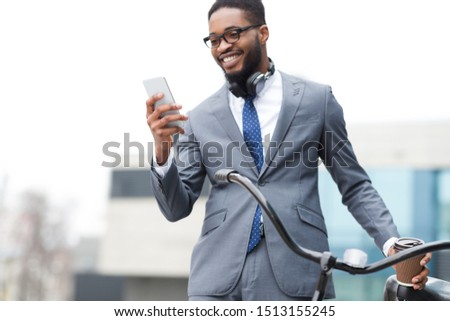 Happy afro entrepreneur using phone, standing with bicycle near office building
