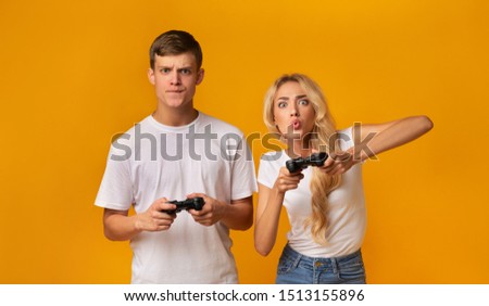 Young guy and girl emotionally playing video game with joysticks, grimacing in excitement over yellow background. Panorama