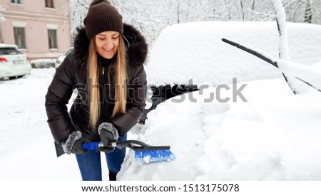Close up of young smiling blond woman in brown coat and jeans trying to clean up snow covered car by brush after a blizzard