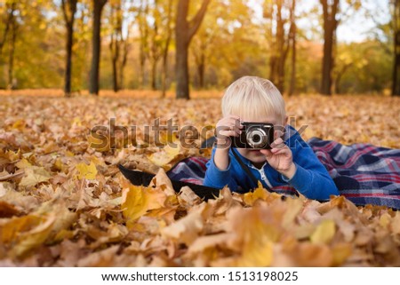 Little blond boy with camera takes pictures. Lying on a plaid, yellow autumn leaves. Fall day