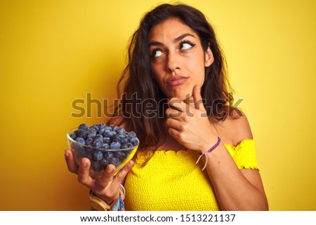 Young beautiful woman holding bowl with blueberries standing over isolated yellow background serious face thinking about question, very confused idea