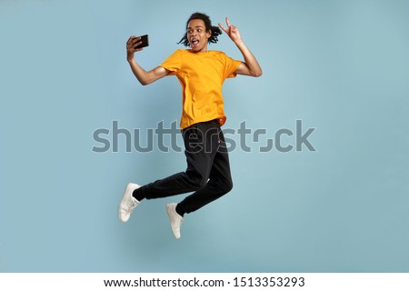 Full length side profile body size photo funky funny young African-American man jump high, show v-sign gesture, make take selfie, wears orange outfit, enjoy a nice day. Isolated blue background
